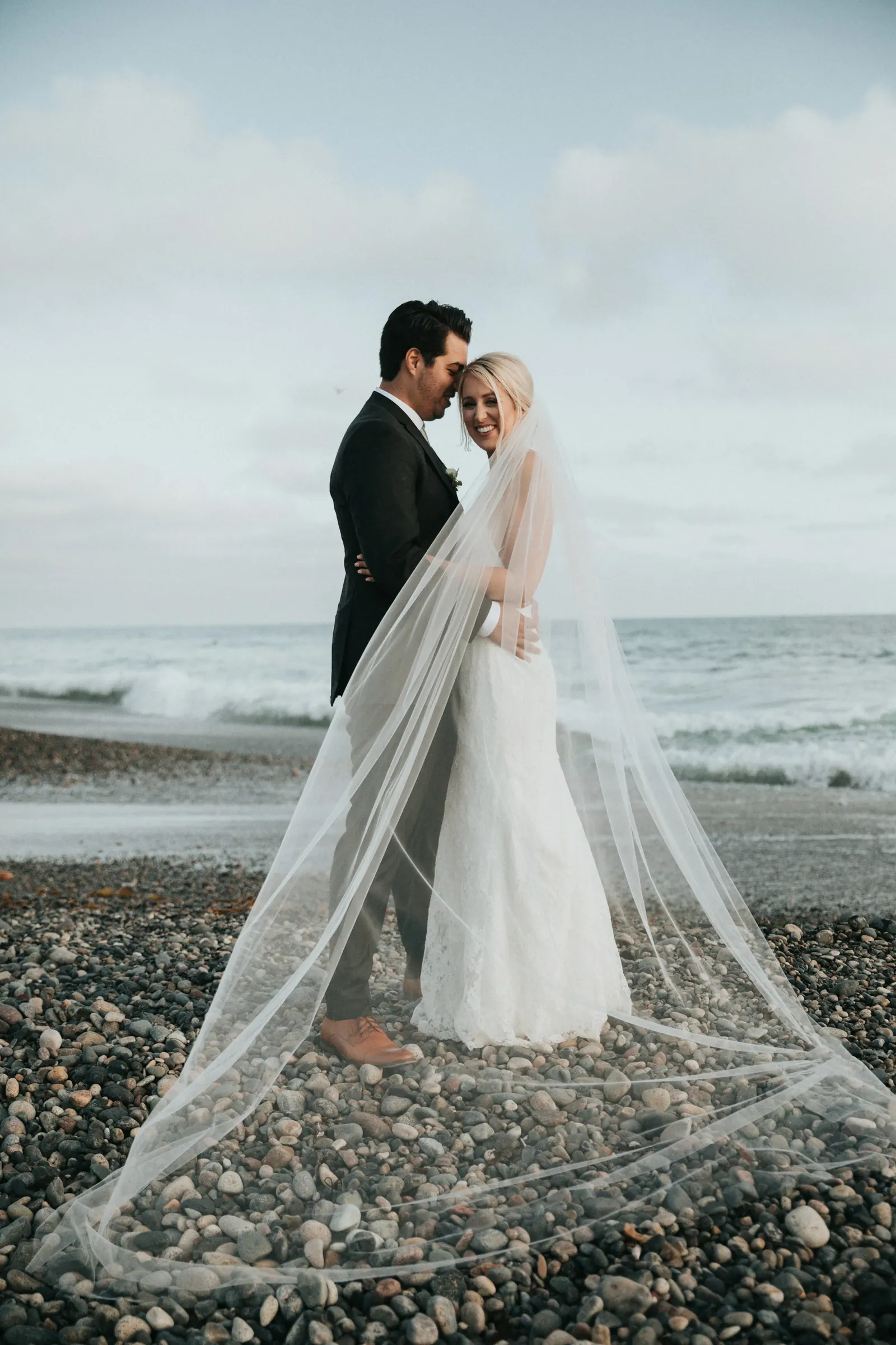Bride and groom on a pebble beach with a cathedral-length veil